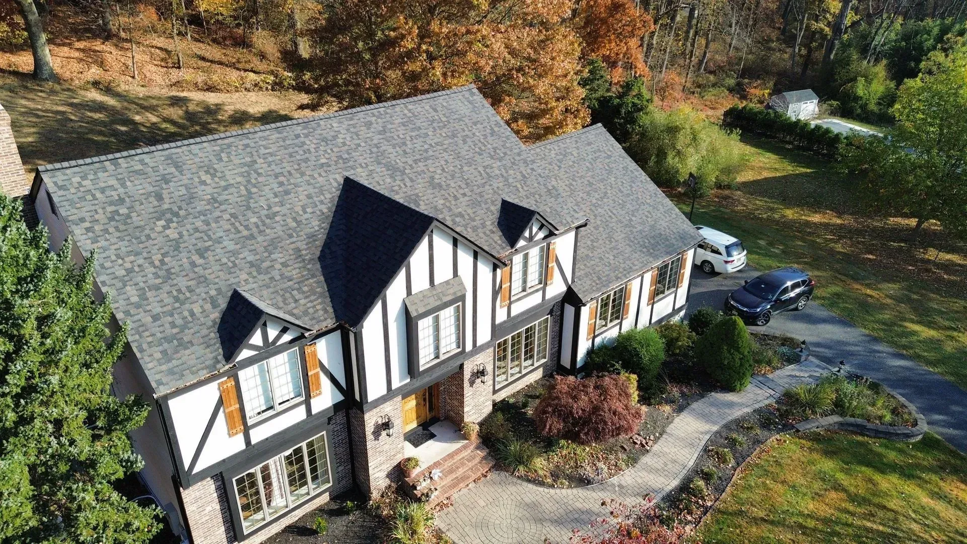Two-story Tudor home with gray roof, black trim, and a curved driveway, surrounded by fall foliage.