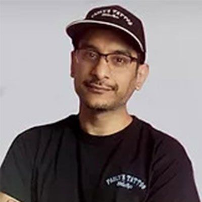 A man wearing a hat and glasses is standing in front of a white wall.