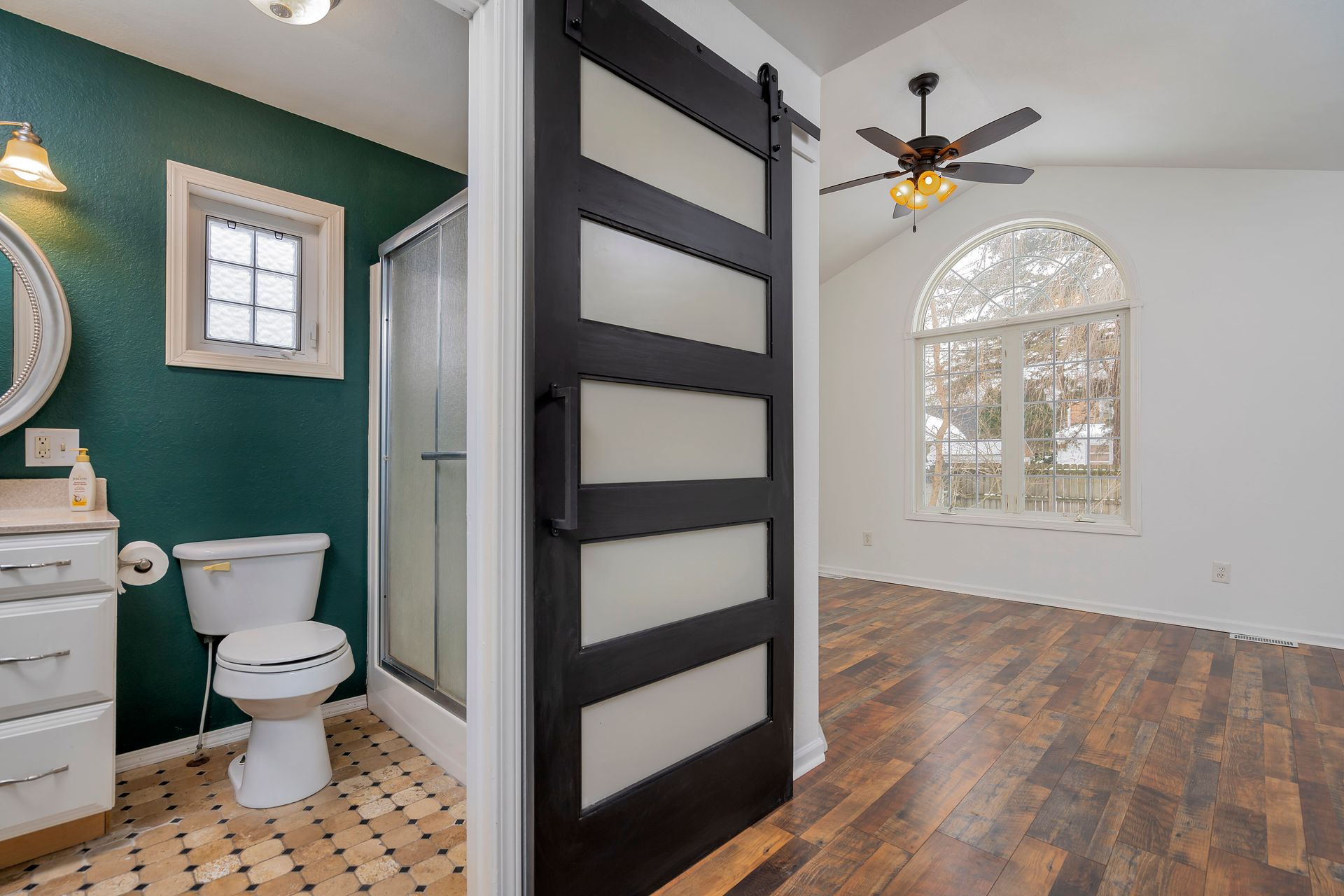 Bathroom and adjacent room with black ladder divider, green walls, white vanity, and arched window in hardwood room