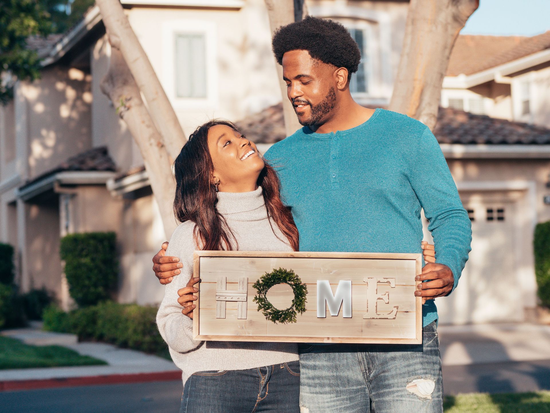 Couple holding a wooden “HOME” sign in front of a house, smiling at each other