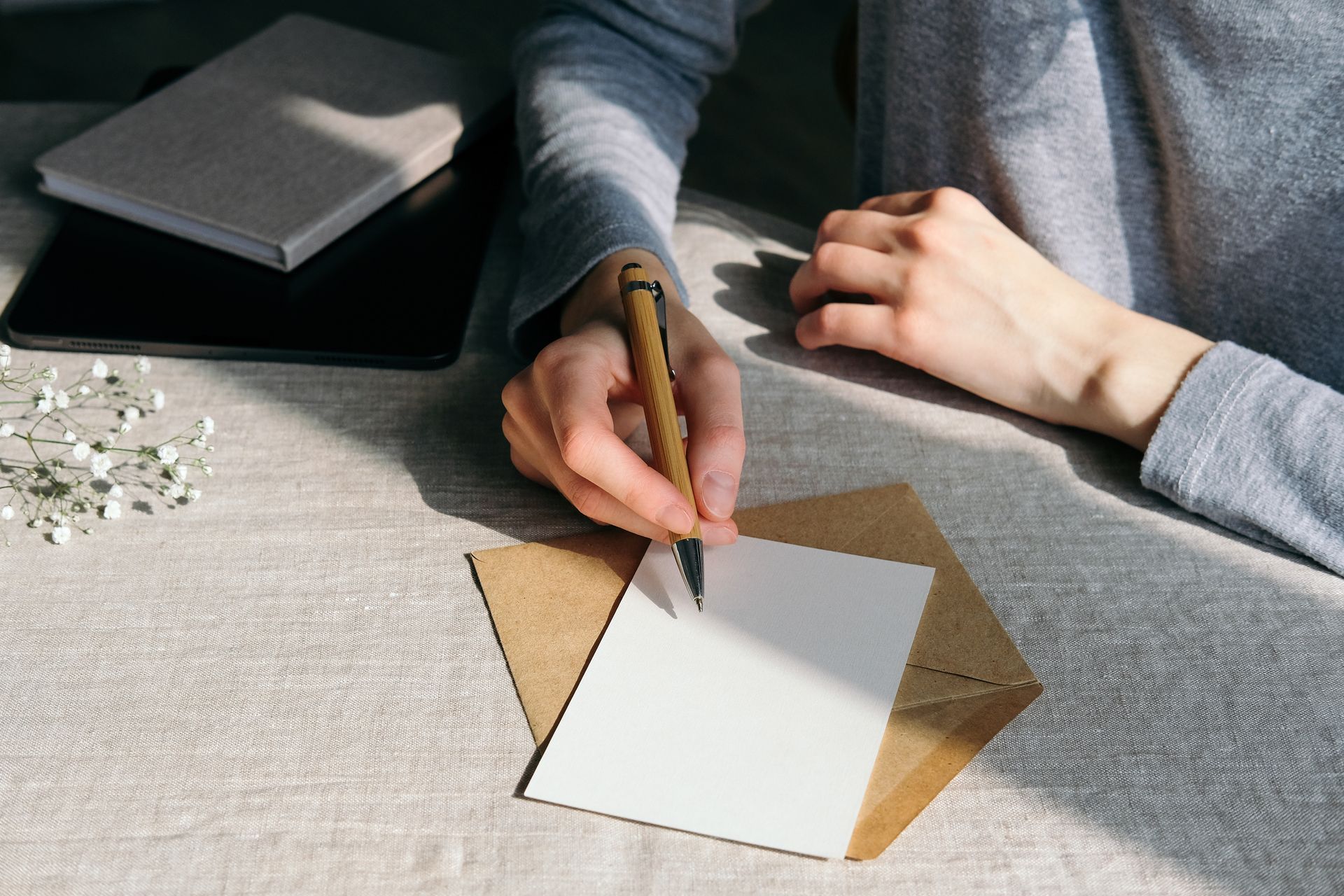 Person writing on a blank card at a sunlit desk with a notebook and envelope nearby