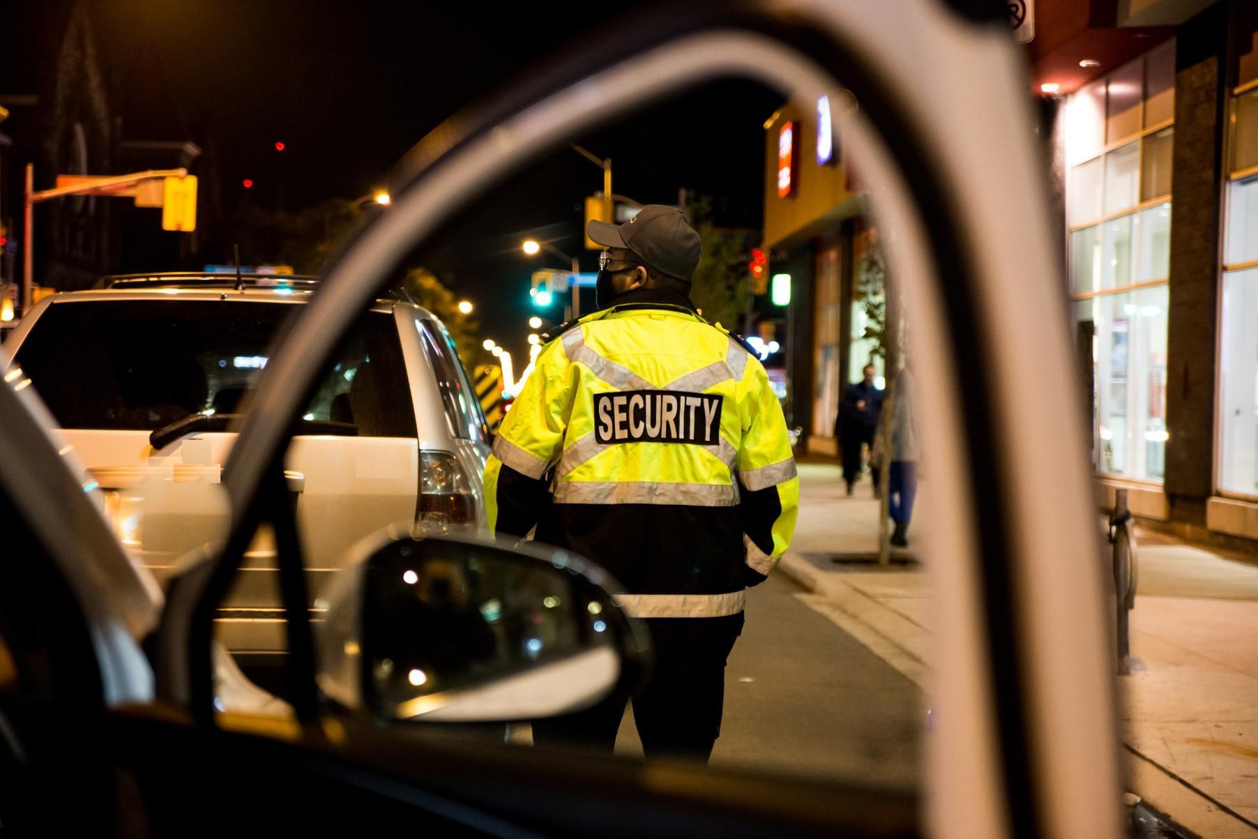 Security guard in a yellow jacket stands on a dimly lit street at night, seen from inside a car.