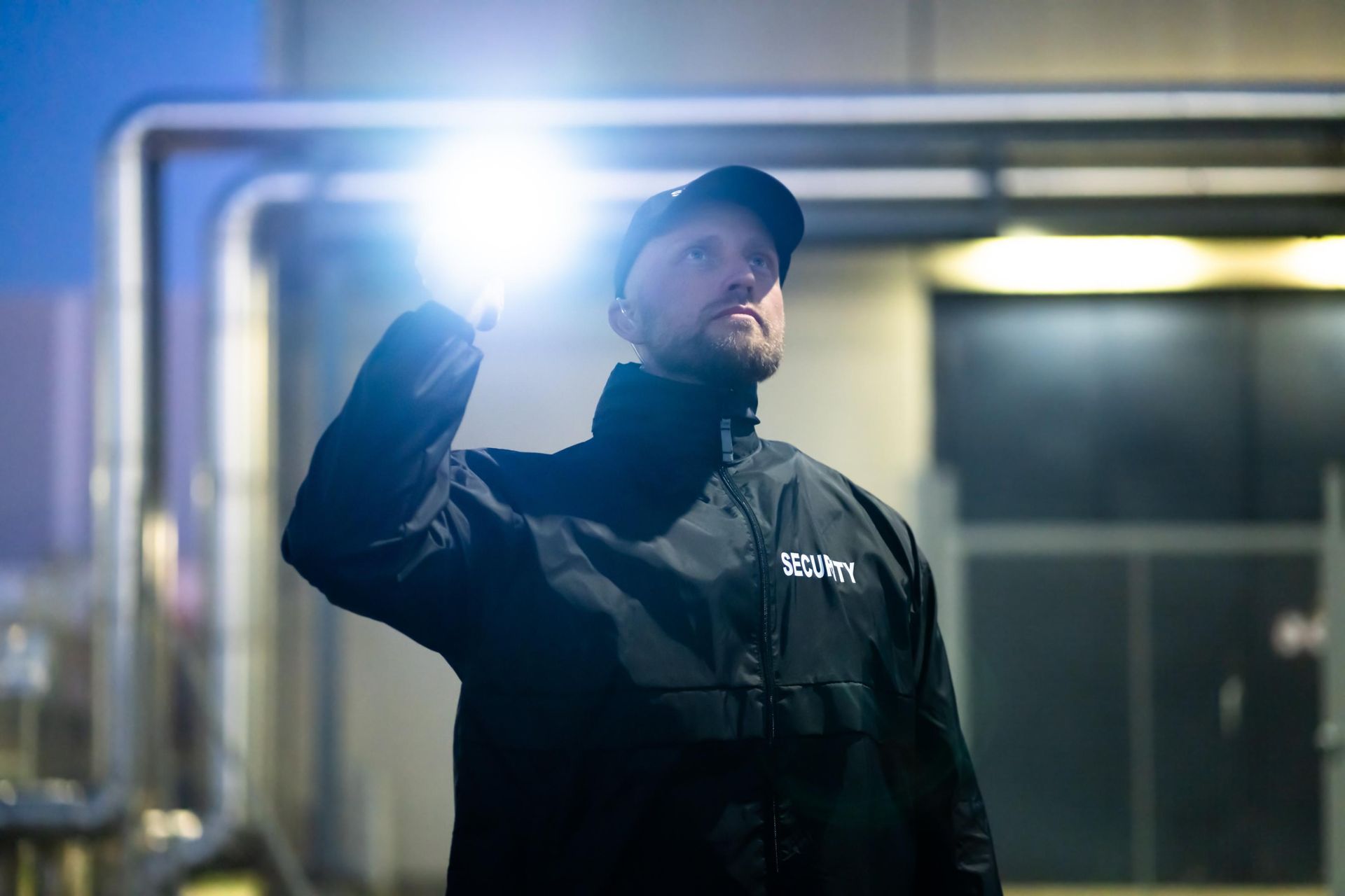Person in dark jacket and cap holds up a bright flashlight at night outside a building