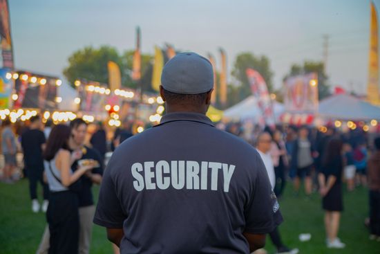 Security guard in dark shirt at outdoor event with crowd and lights in background
