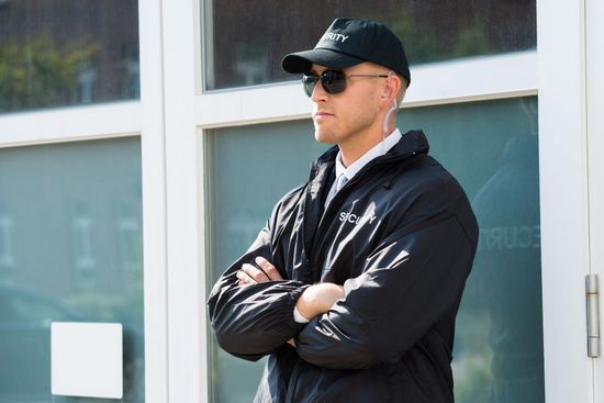 Person in a black cap and jacket stands with arms crossed outside a building.