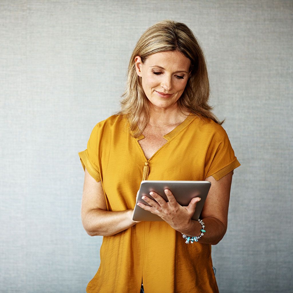 Donna bionda con una maglietta gialla, che guarda un tablet, sorridente, sfondo grigio.