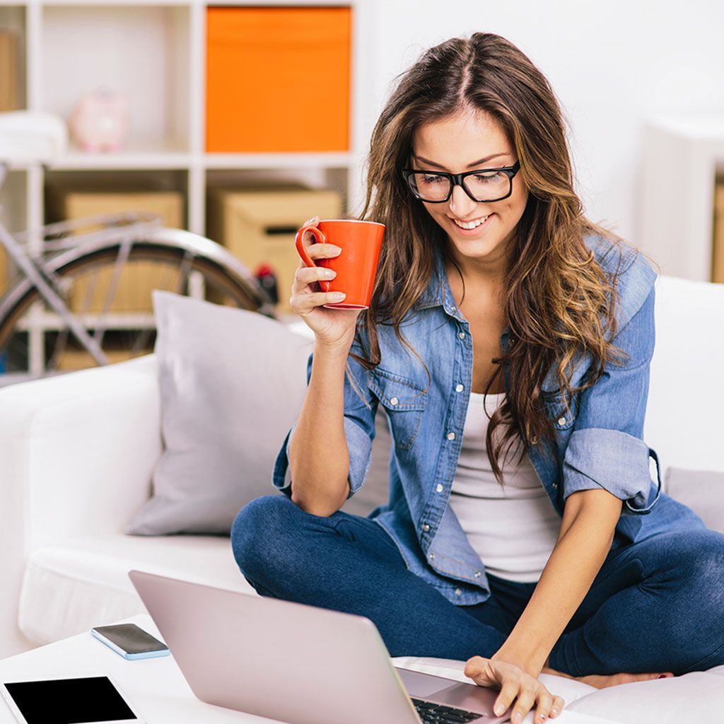 Donna sul divano con il portatile, beve caffè, sorride. Camicia di jeans, occhiali, ambiente domestico.