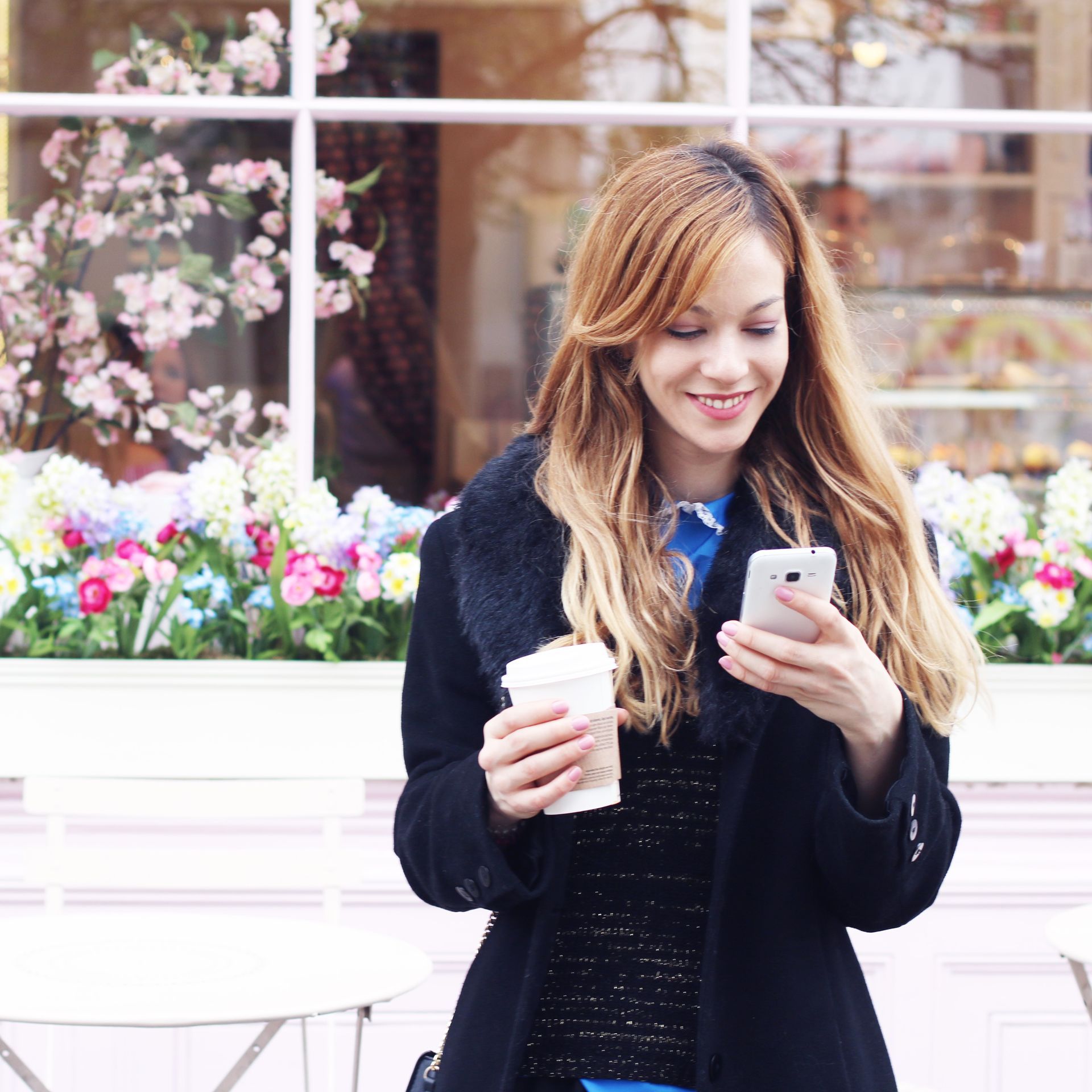 Donna in cappotto, sorridente, che guarda il telefono, con in mano un caffè, davanti a una finestra piena di fiori.