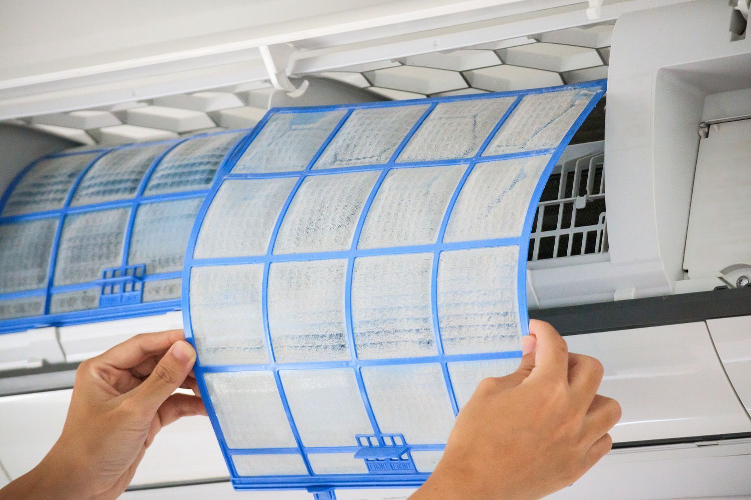 Hands removing a dust-covered air conditioner filter for cleaning.
