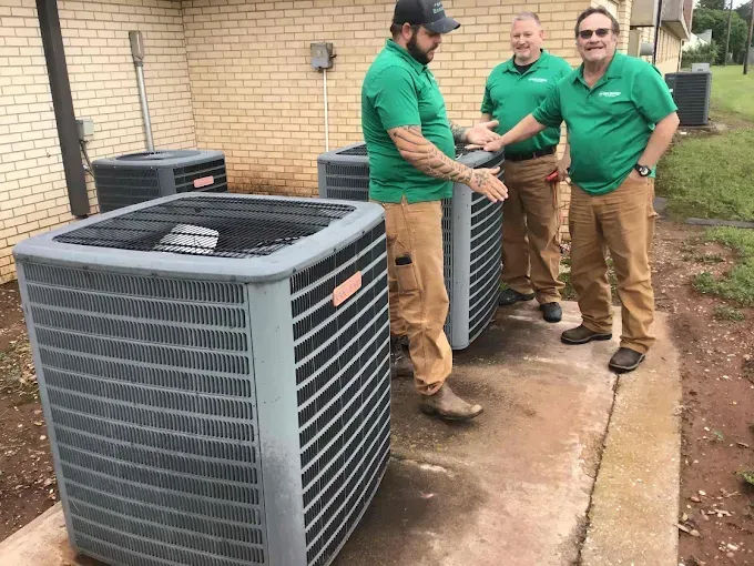 Three technicians in matching green shirts and tan work pants stand outside, posing next to several HVAC condenser units.