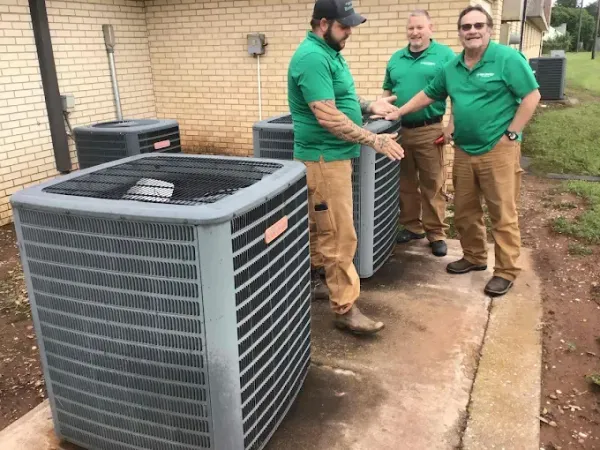 Three technicians in green shirts stand on a concrete pad next to several large, gray outdoor HVAC units.