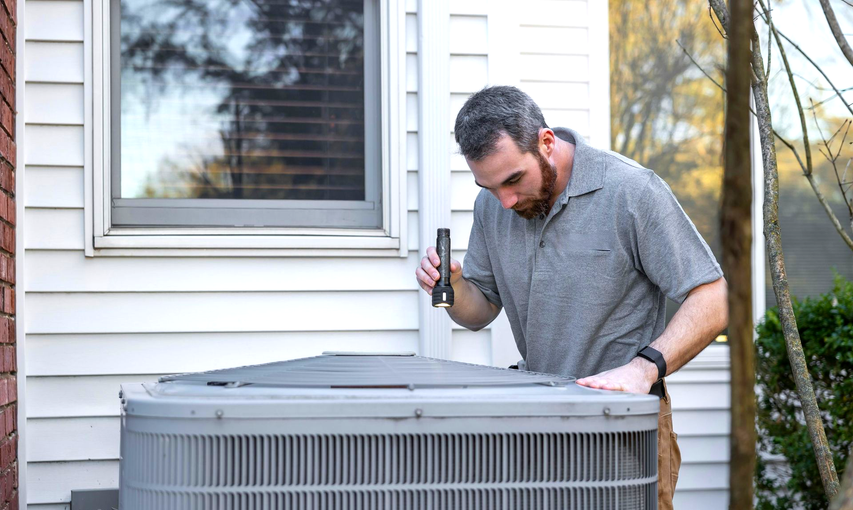 A technician inspects an outdoor air conditioning unit with a flashlight.
