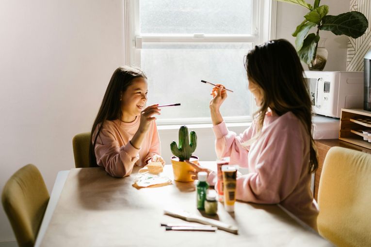 Two people in pink sweaters smile while crafting together at a table near a window, with a small cactus plant between them.