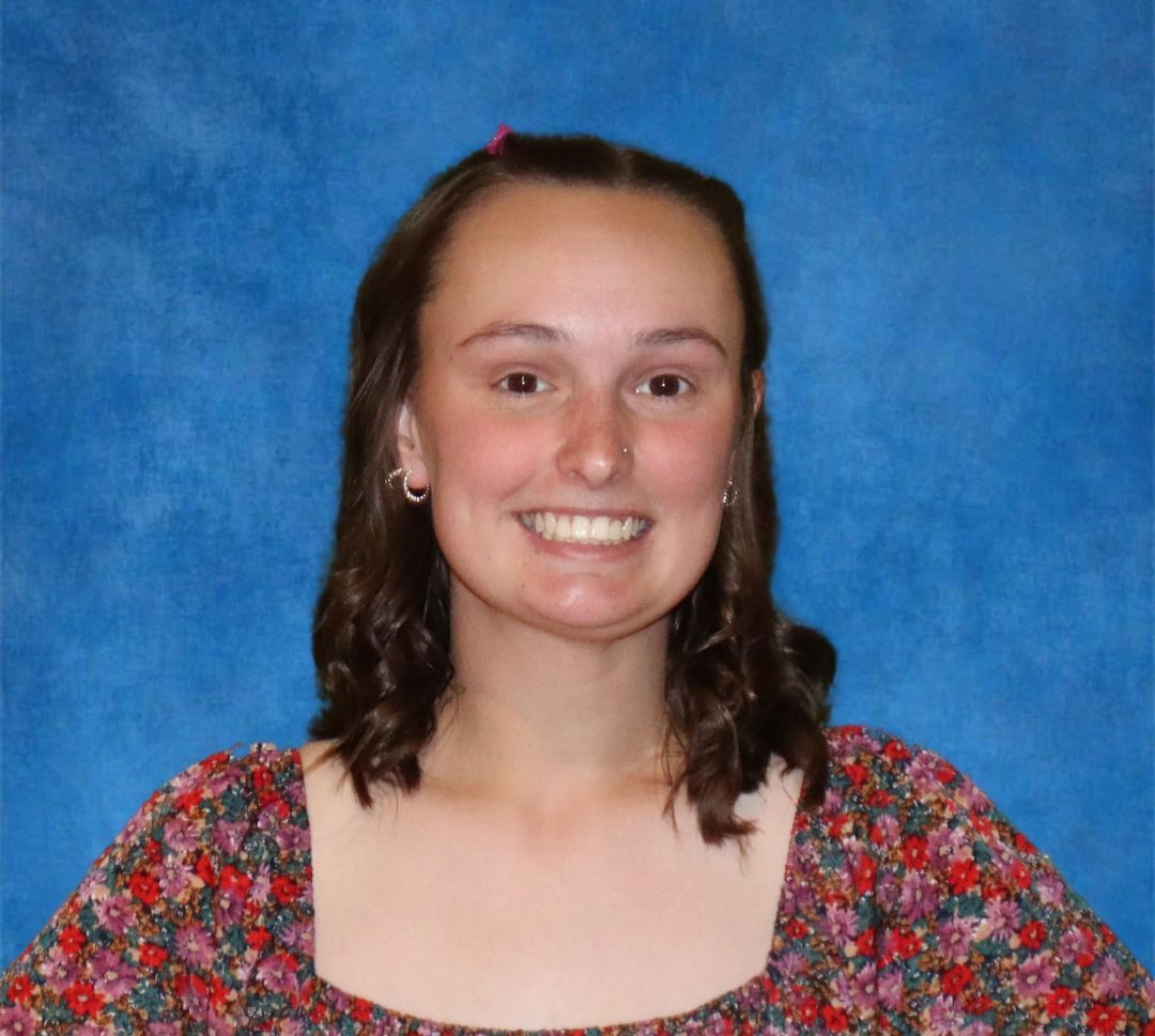 A young woman in a floral dress is smiling in front of a blue background.