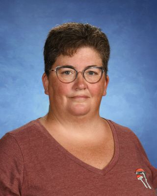 A woman wearing glasses and a red shirt is standing in front of a blue background. This is Christina Tomsett.