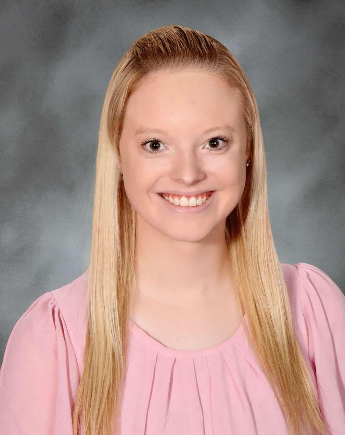 A young woman in a pink shirt is smiling for a picture. This is Ashley Geckle.