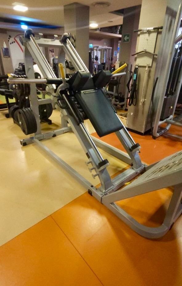 Macchina per leg press angolata in una palestra con pavimento arancione e attrezzature a specchio tutt'intorno.