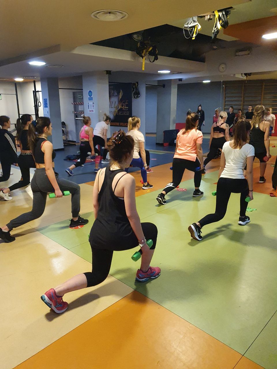 Persone che si allenano in una palestra, eseguendo affondi sincronizzati su un pavimento colorato.