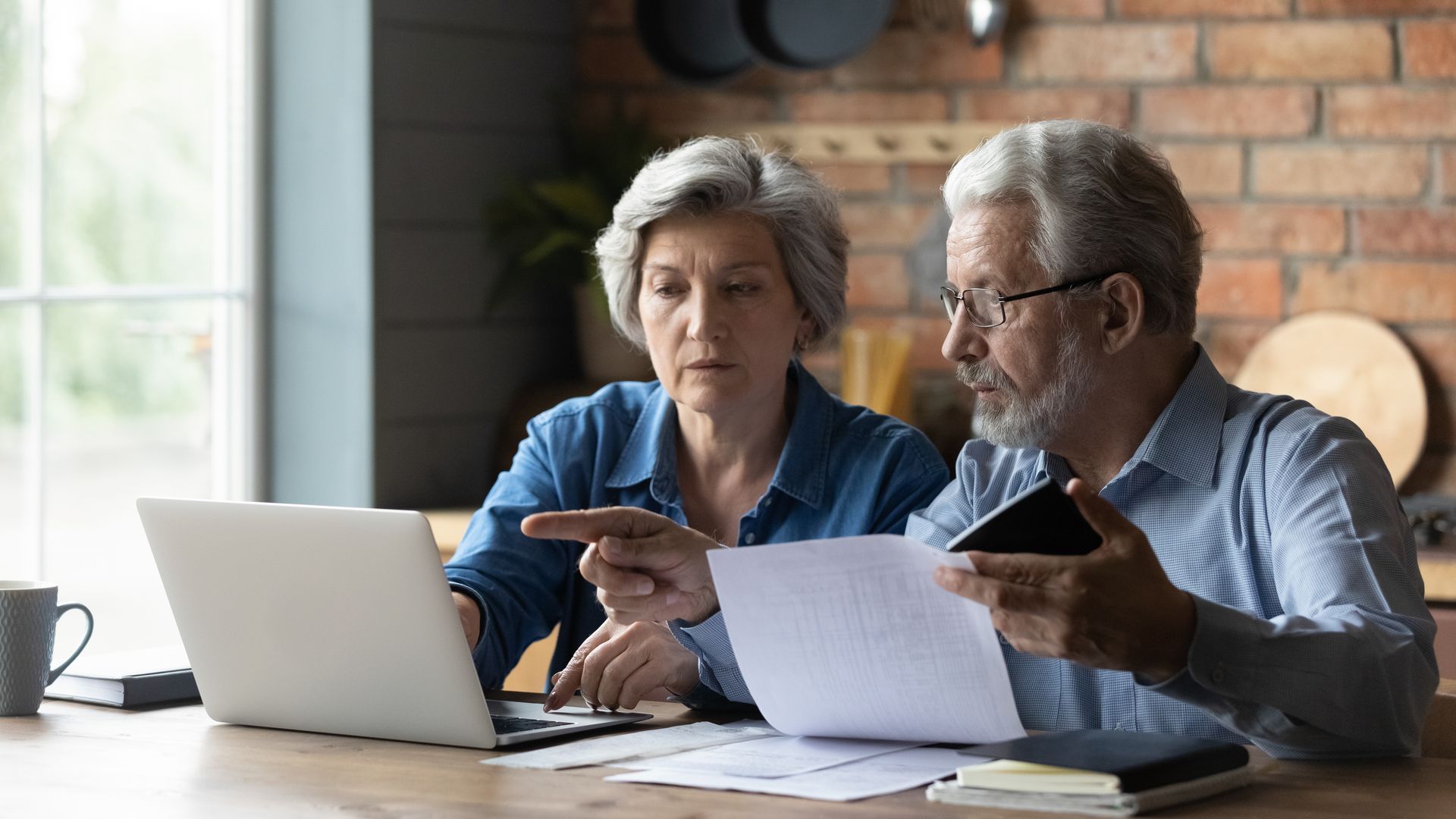Two people reviewing papers beside a laptop at a table in a bright home office