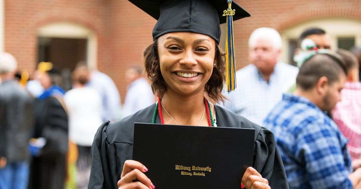 Graduate smiling in cap and gown, holding diploma folder at an outdoor ceremony.