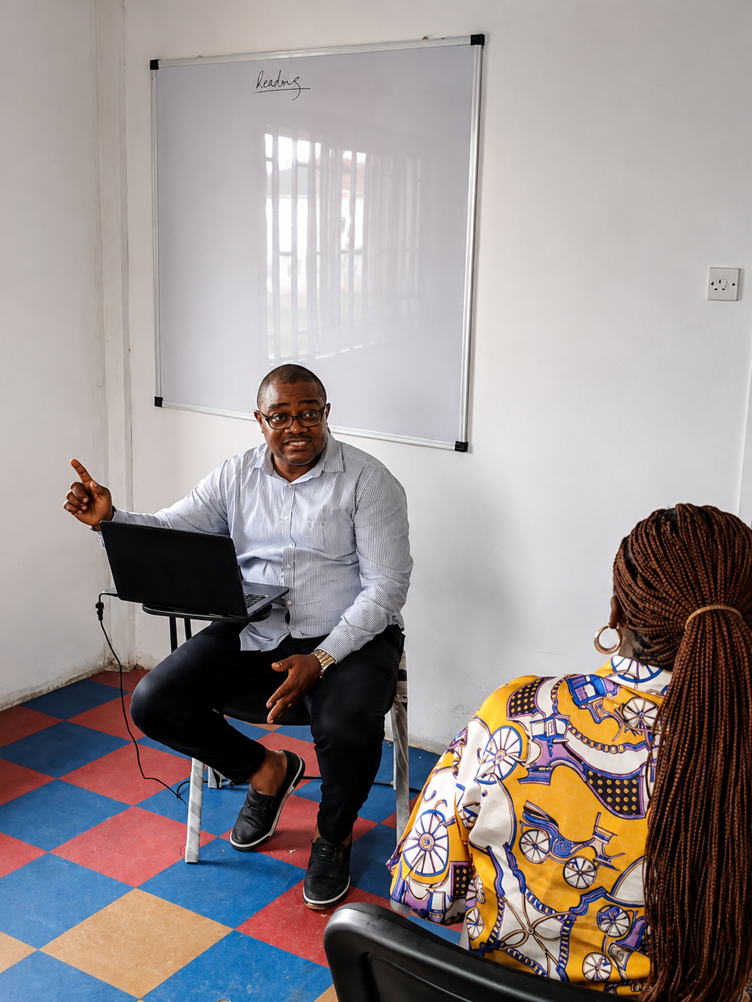 Man speaking beside a laptop in a small meeting room, with a whiteboard on the wall and a seated listener nearby.