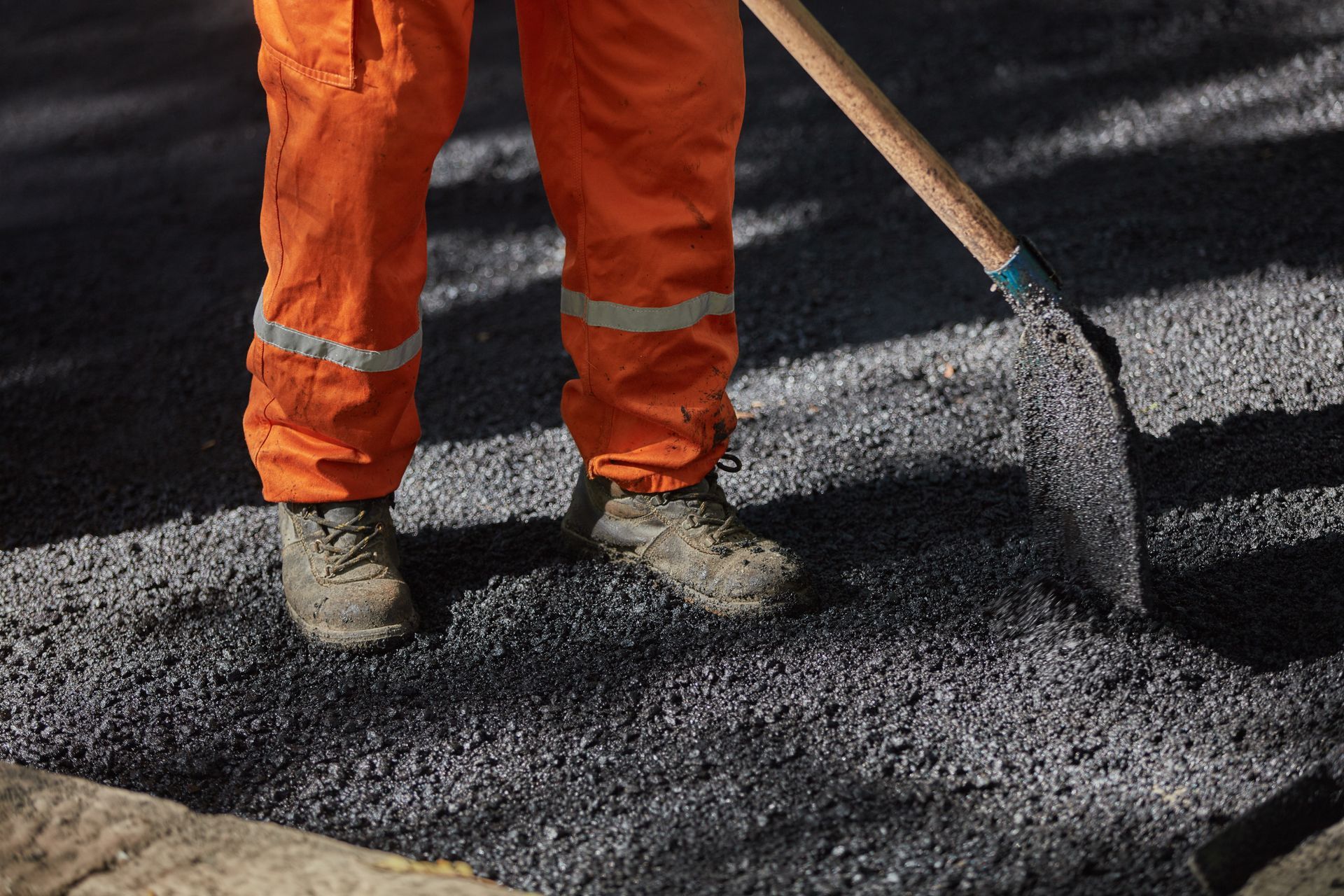 Worker in orange safety pants shoveling fresh black asphalt on a road surface Worker in orange safety pants shoveling fresh black asphalt on a road surface