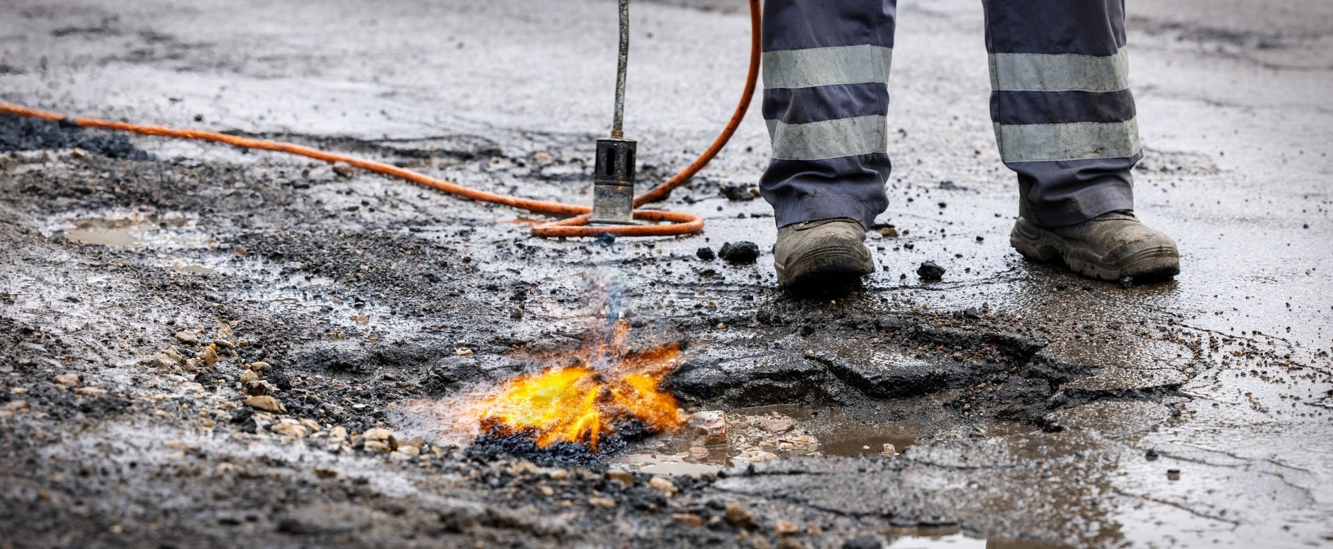 Fire on wet pavement near a person’s boots and a hose, with smoke rising. Fire on wet pavement near a person’s boots and a hose, with smoke rising.