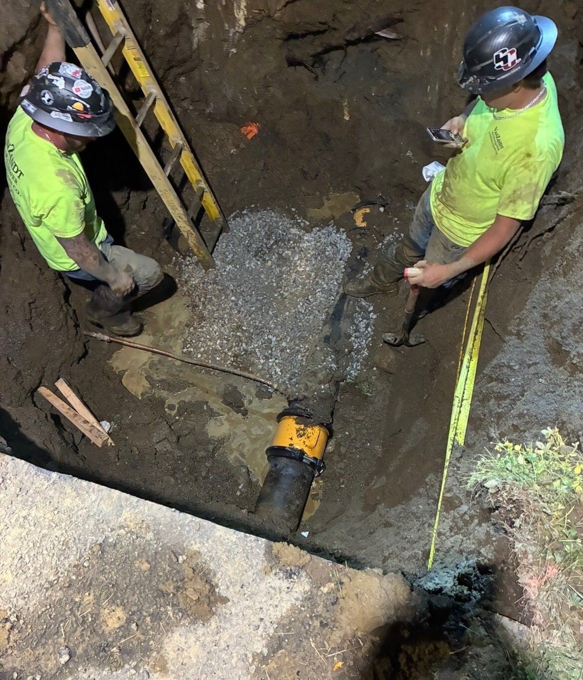 Two workers in hard hats inspect a sewer trench around a pipe opening. Two workers in hard hats inspect a sewer trench around a pipe opening.