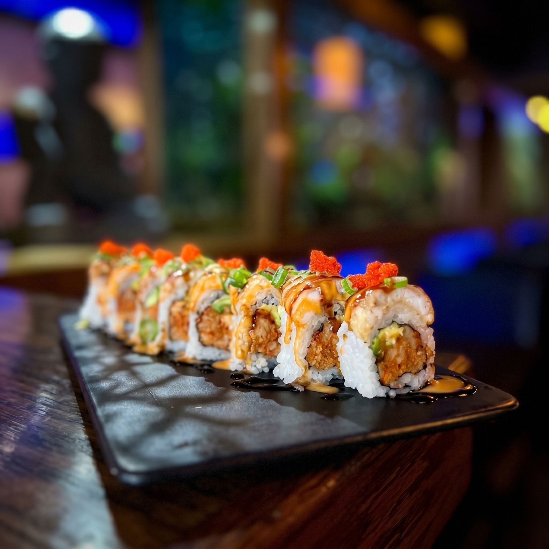 Sushi rolls topped with sauce and garnish on a black plate in a dimly lit restaurant