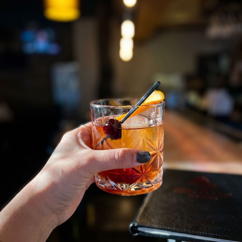 Hand holding a whiskey cocktail with orange peel in a dimly lit bar