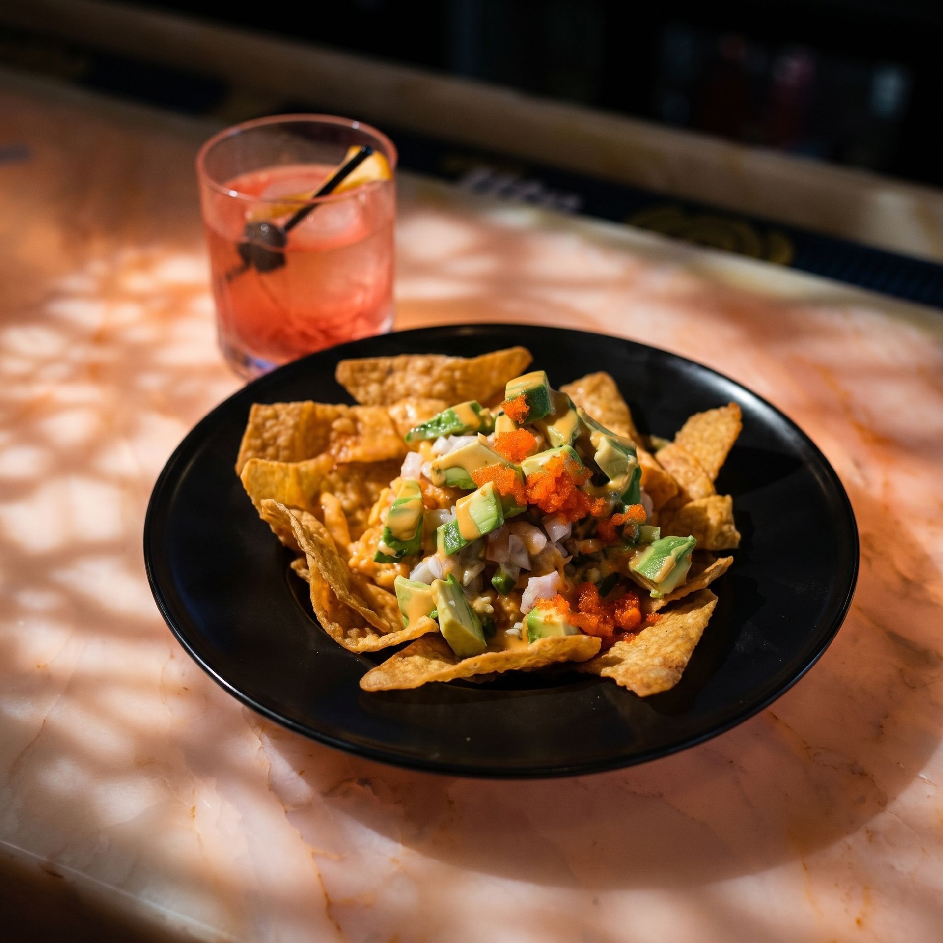 Black bowl of loaded nachos with salsa and guacamole beside a pink cocktail on a table