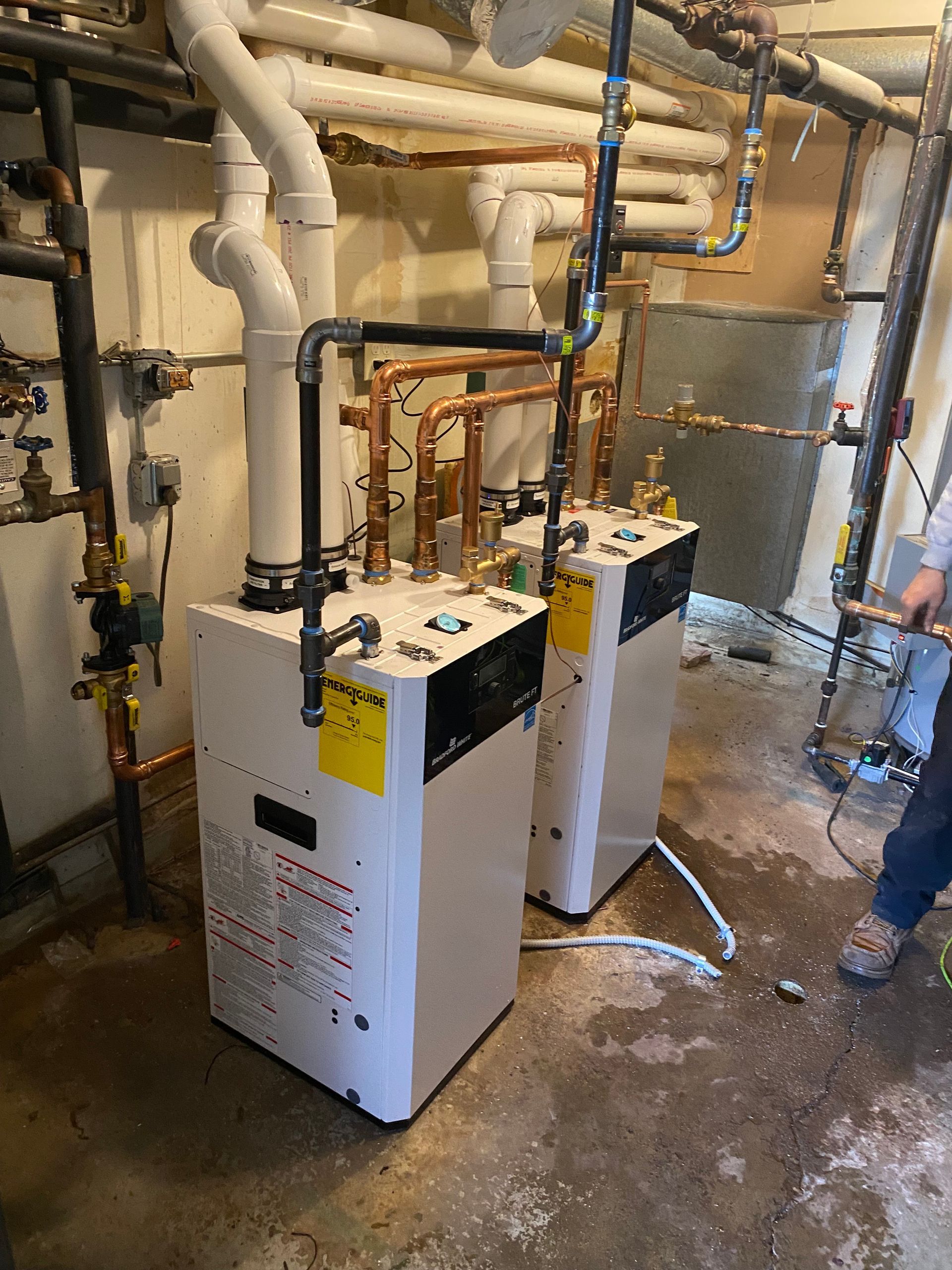 Two white boiler units in a basement utility room with exposed pipes and concrete floor.