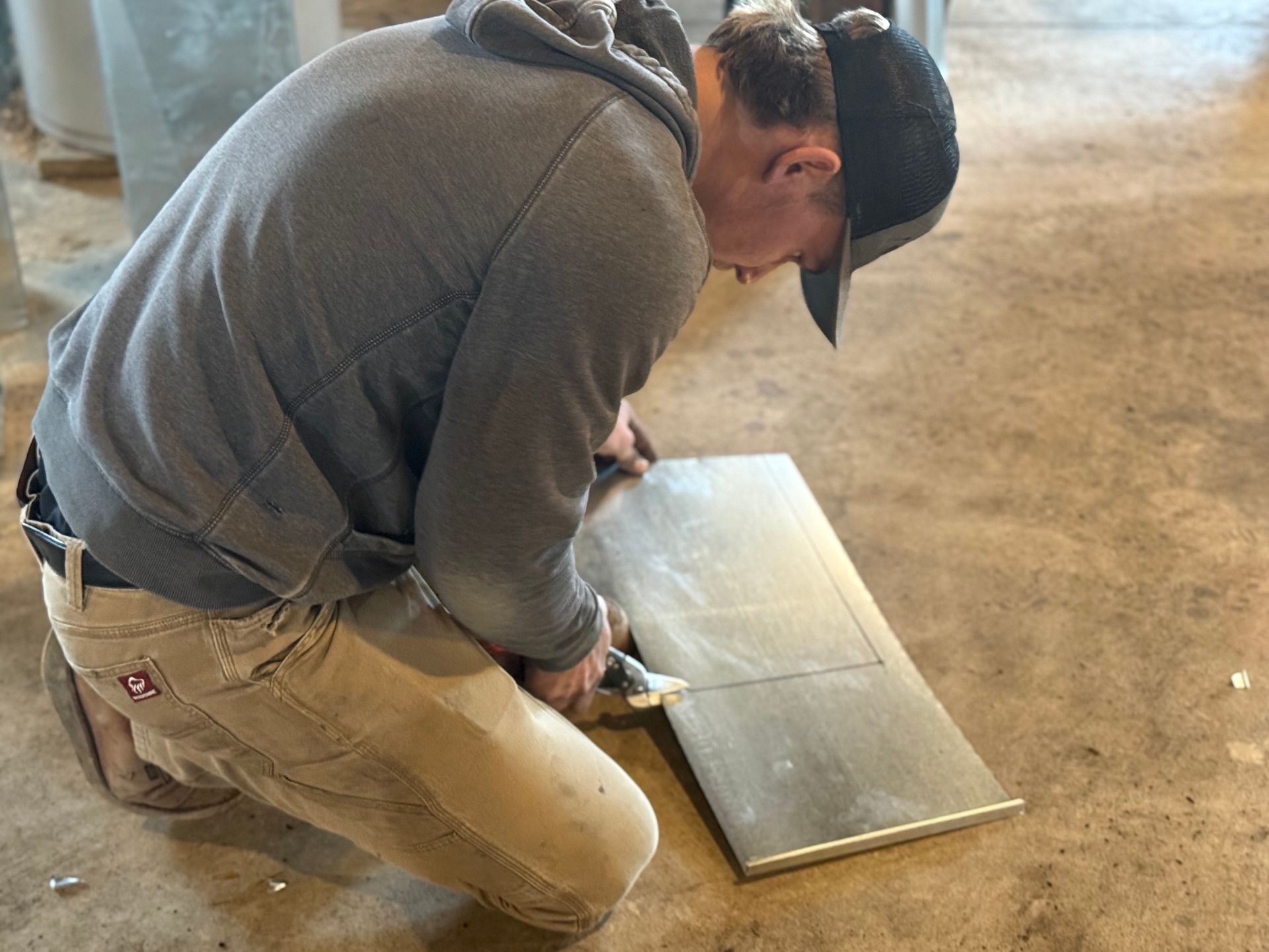 Person kneeling on a concrete floor, using a tool to cut a gray metal panel