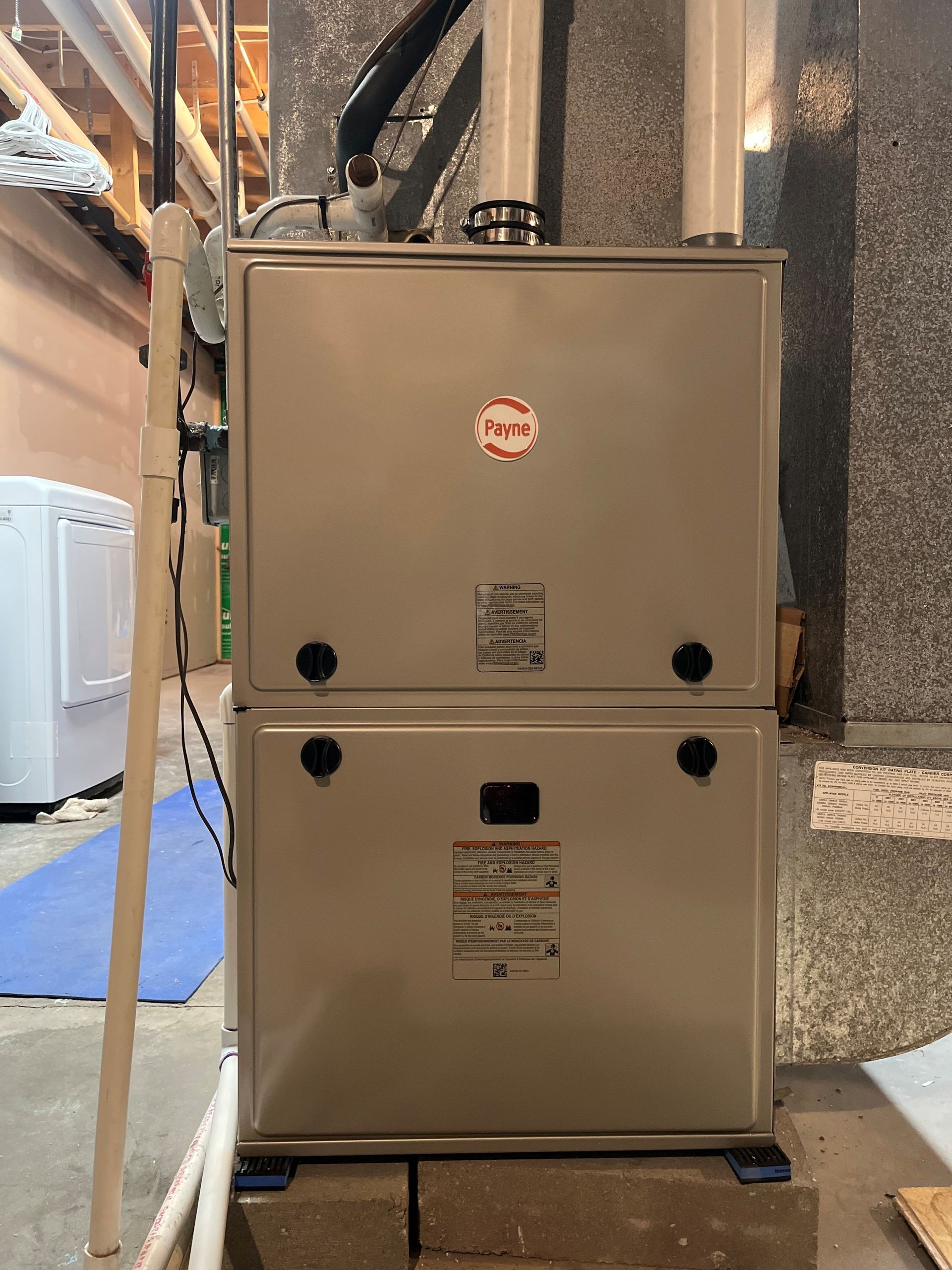 Two stacked beige furnace units in a basement utility room with visible ducts and pipes.