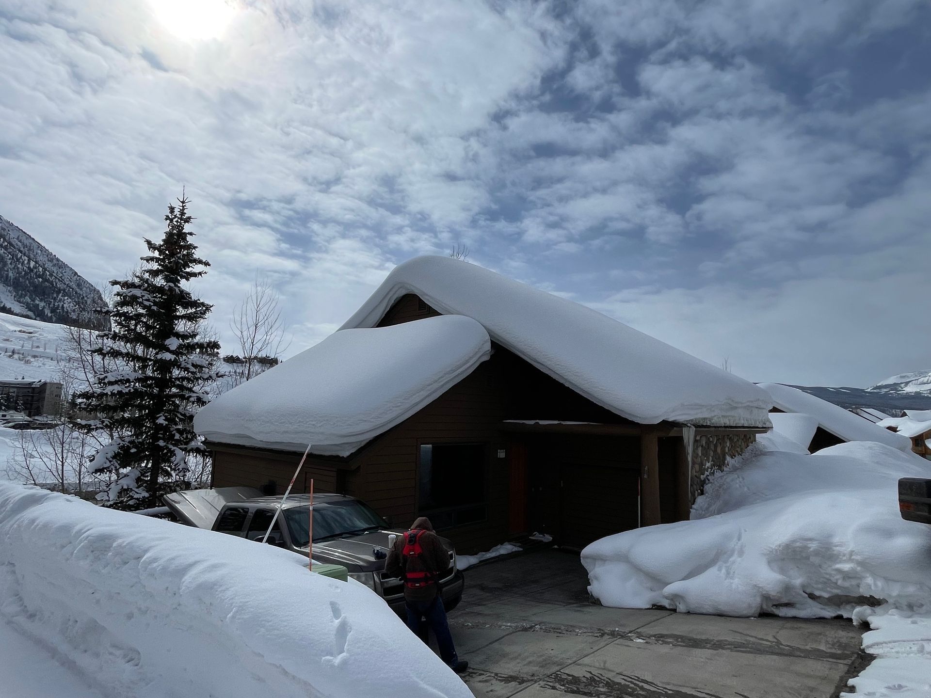 Snowy scene of a wooden house with a snow-covered roof, driveway, and large snowbanks.