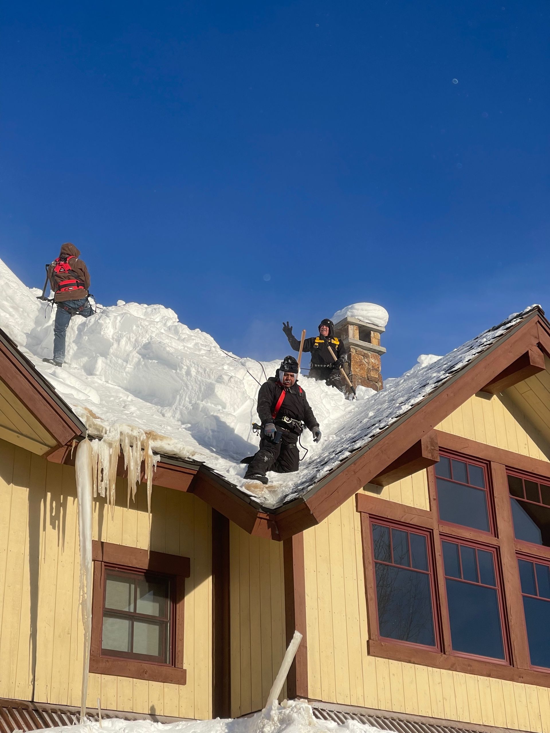 People clearing snow from a roof. Yellow house, snowy roof, clear blue sky, icicles.