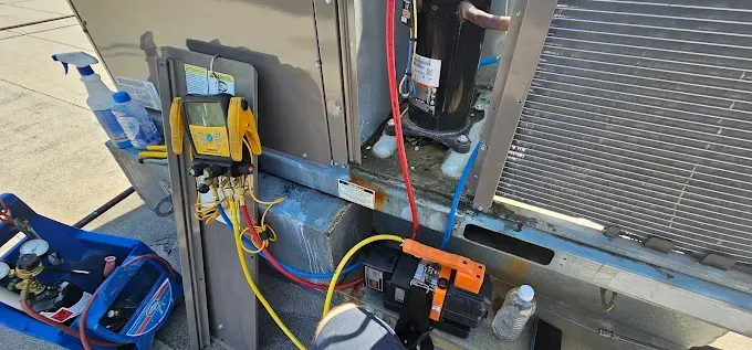 HVAC technician working on an air conditioning unit. Yellow and red hoses connected to gauges. Unit is outdoors.
