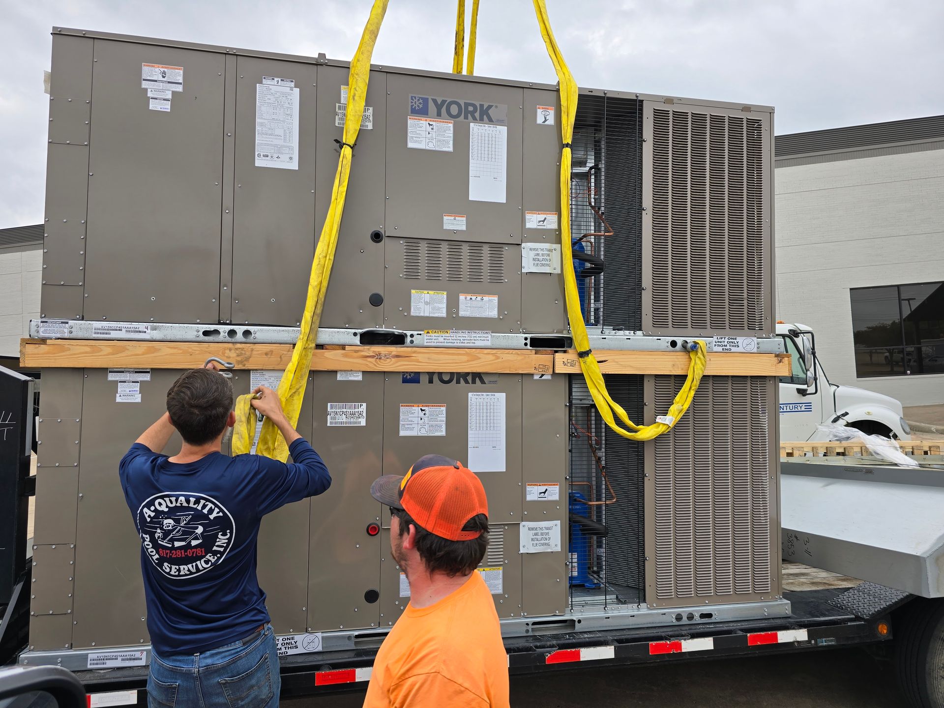 Two HVAC units stacked on a trailer, lifted by yellow straps, with two workers inspecting.