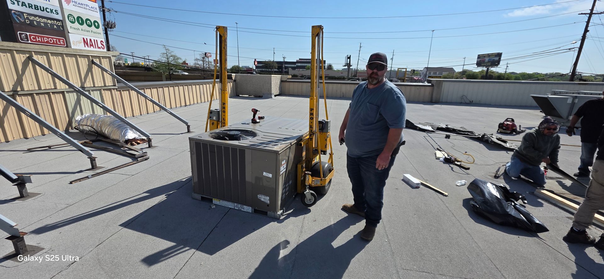 A person working on machinery outdoors, wearing a cap and holding tools near a large fan.