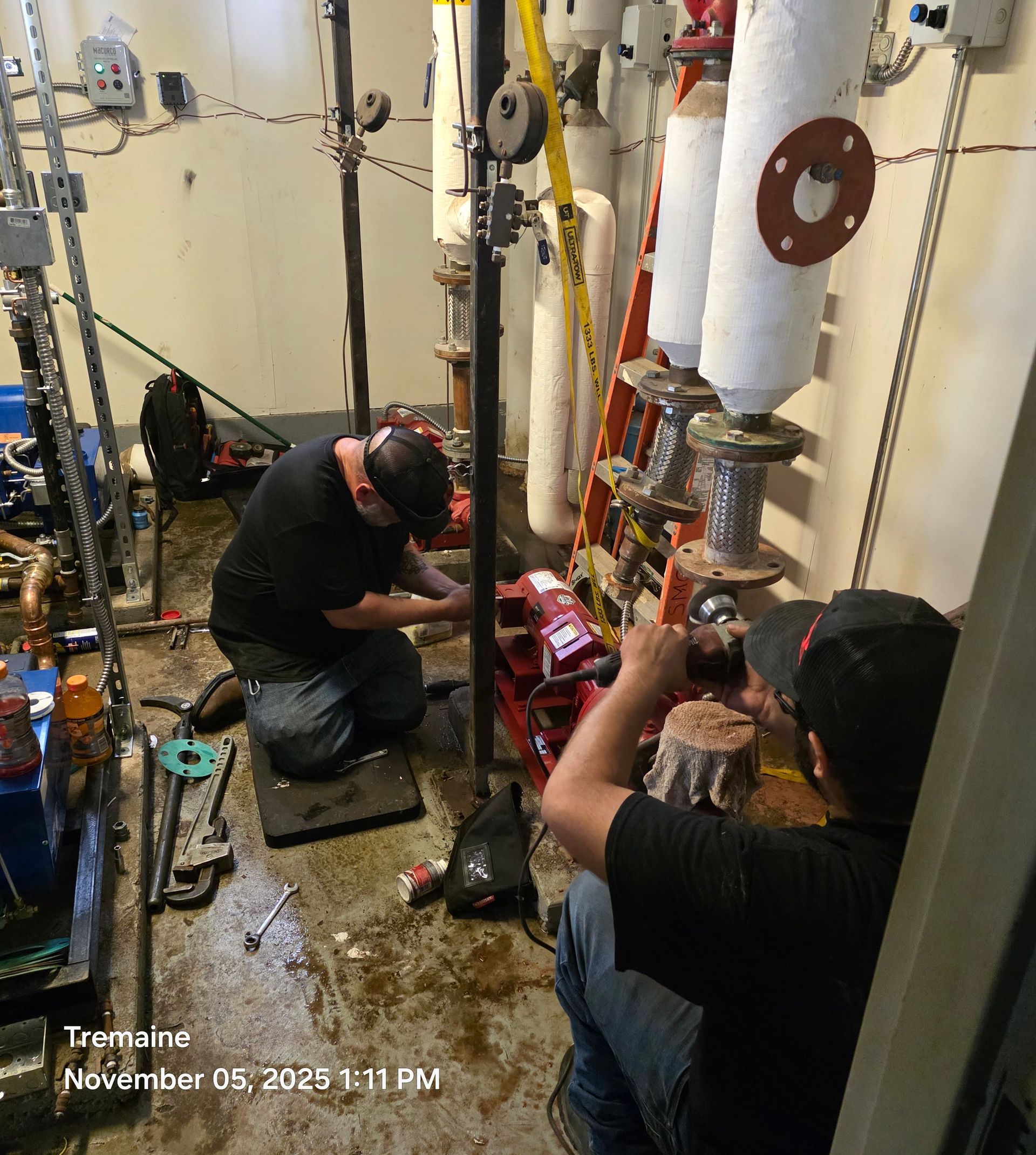 Two men working on pipes in an industrial setting, one kneeling, one seated.