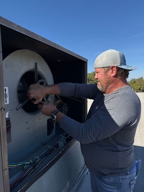 A person working on machinery outdoors, wearing a cap and holding tools near a large fan.