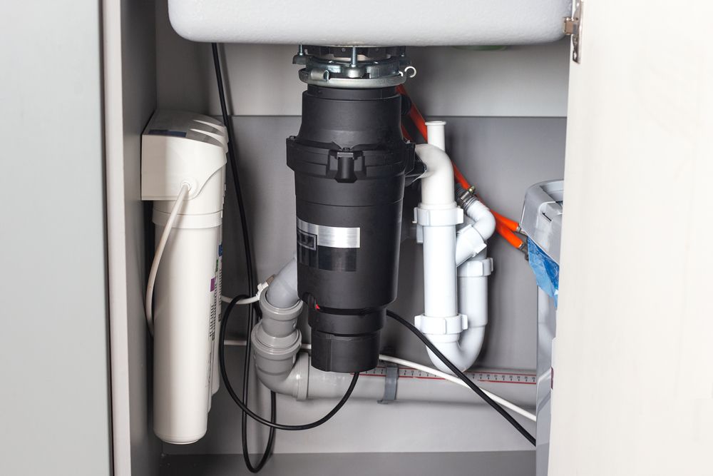 Undersink plumbing with garbage disposal, water filter, and drainpipes. Gray cabinet, white sink, black disposal.