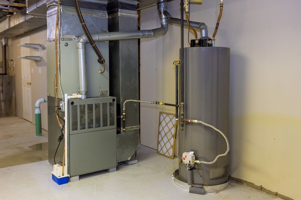 Furnace and water heater in a basement. Grey appliances with silver ductwork and pipes against a light-colored wall.