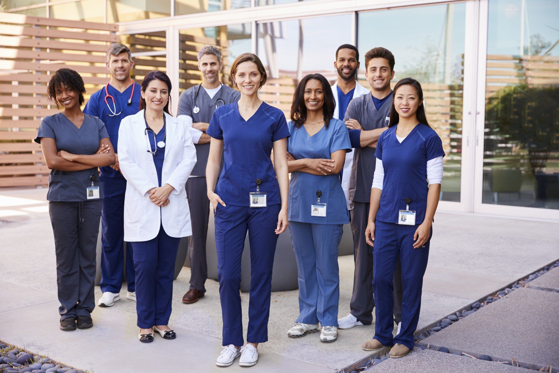 Group of healthcare professionals in blue scrubs and white coats, smiling outside a modern building.