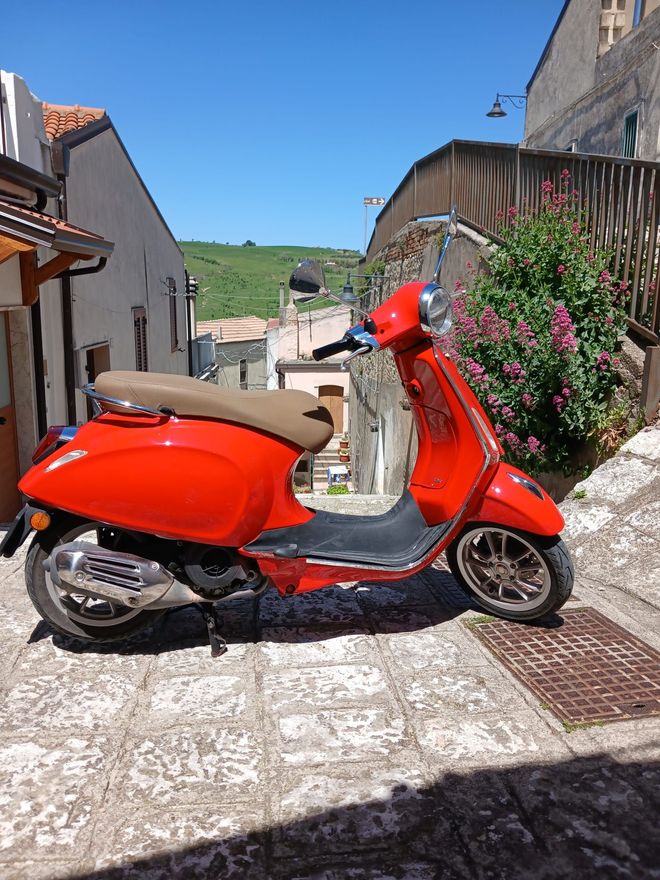 Red scooter parked on a stone street between buildings under a bright blue sky.