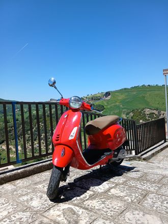 Red scooter parked on a stone terrace overlooking green hills under a clear blue sky.