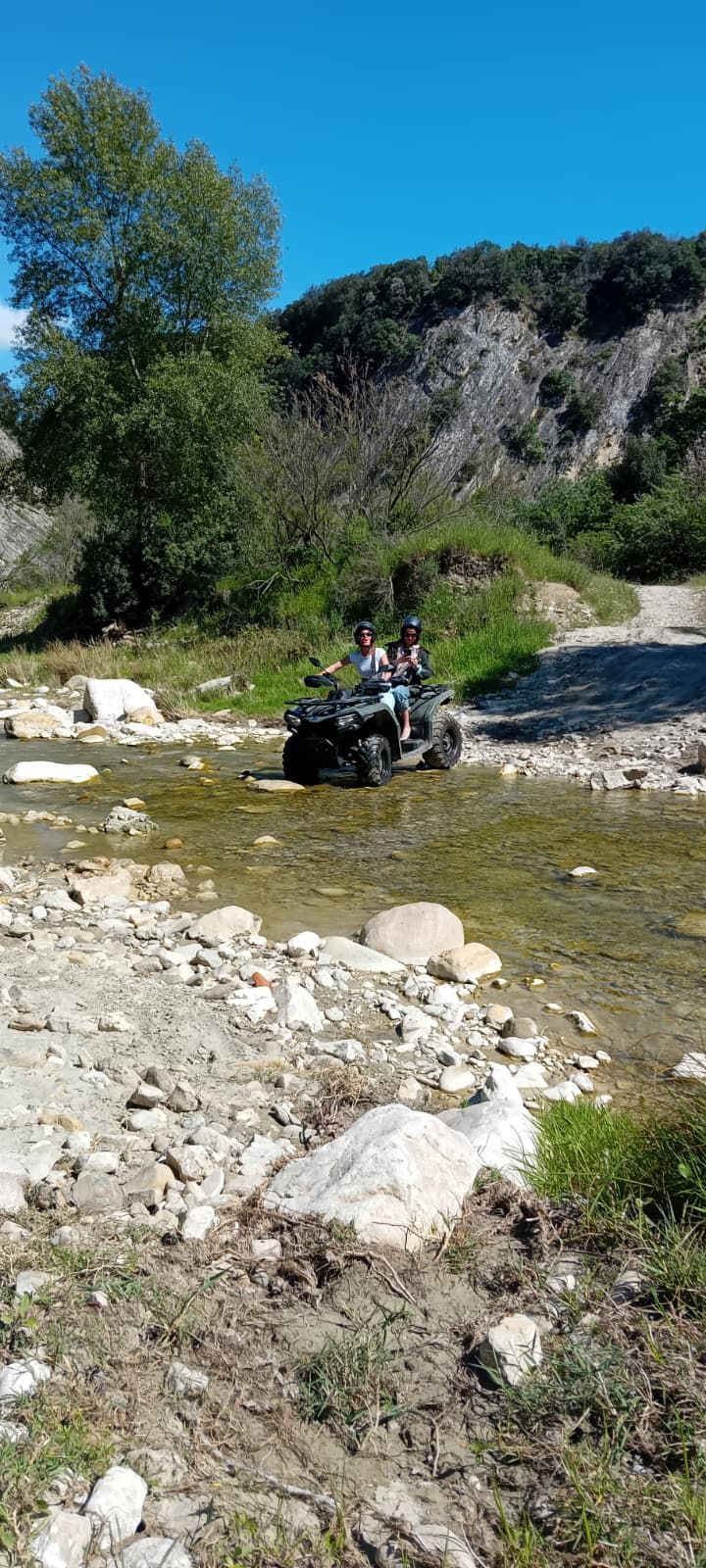 ATV with riders crossing a shallow rocky stream in a sunny mountain valley