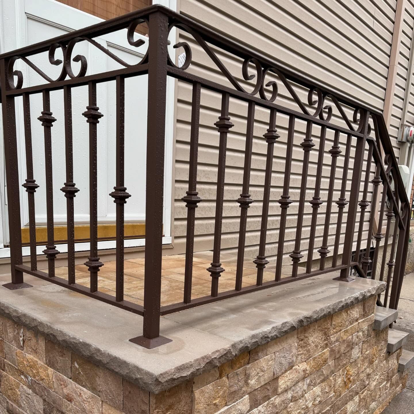 Brown metal railings with decorative scrolls and spindles installed on a stone-masonry porch next to a house.