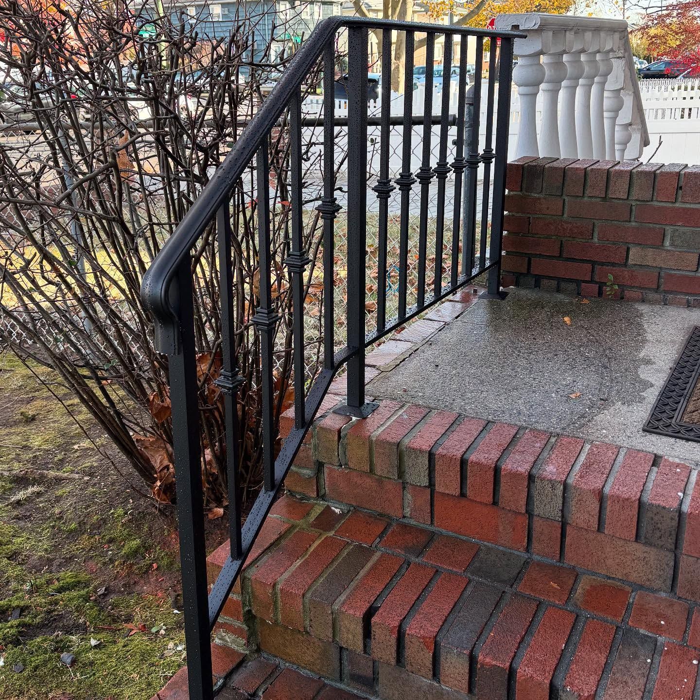 Black metal handrail on brick steps leading to a white porch railing beside a bare bush.