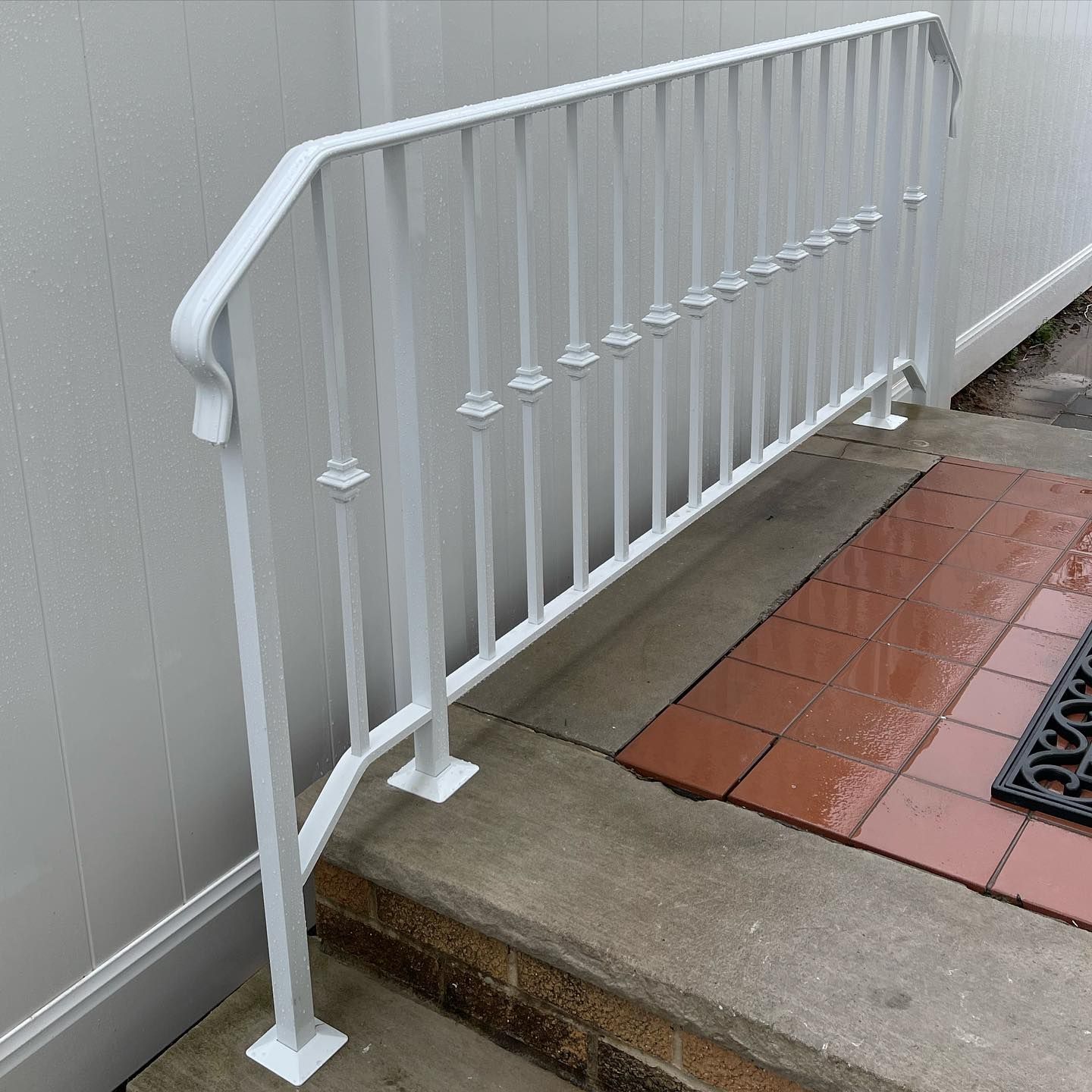 A white metal railing sits on concrete steps against a white vinyl fence and a brick patio.